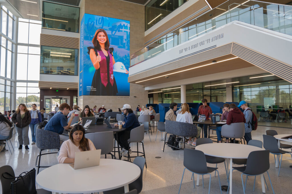 Many students sitting at tables in front of a large screen