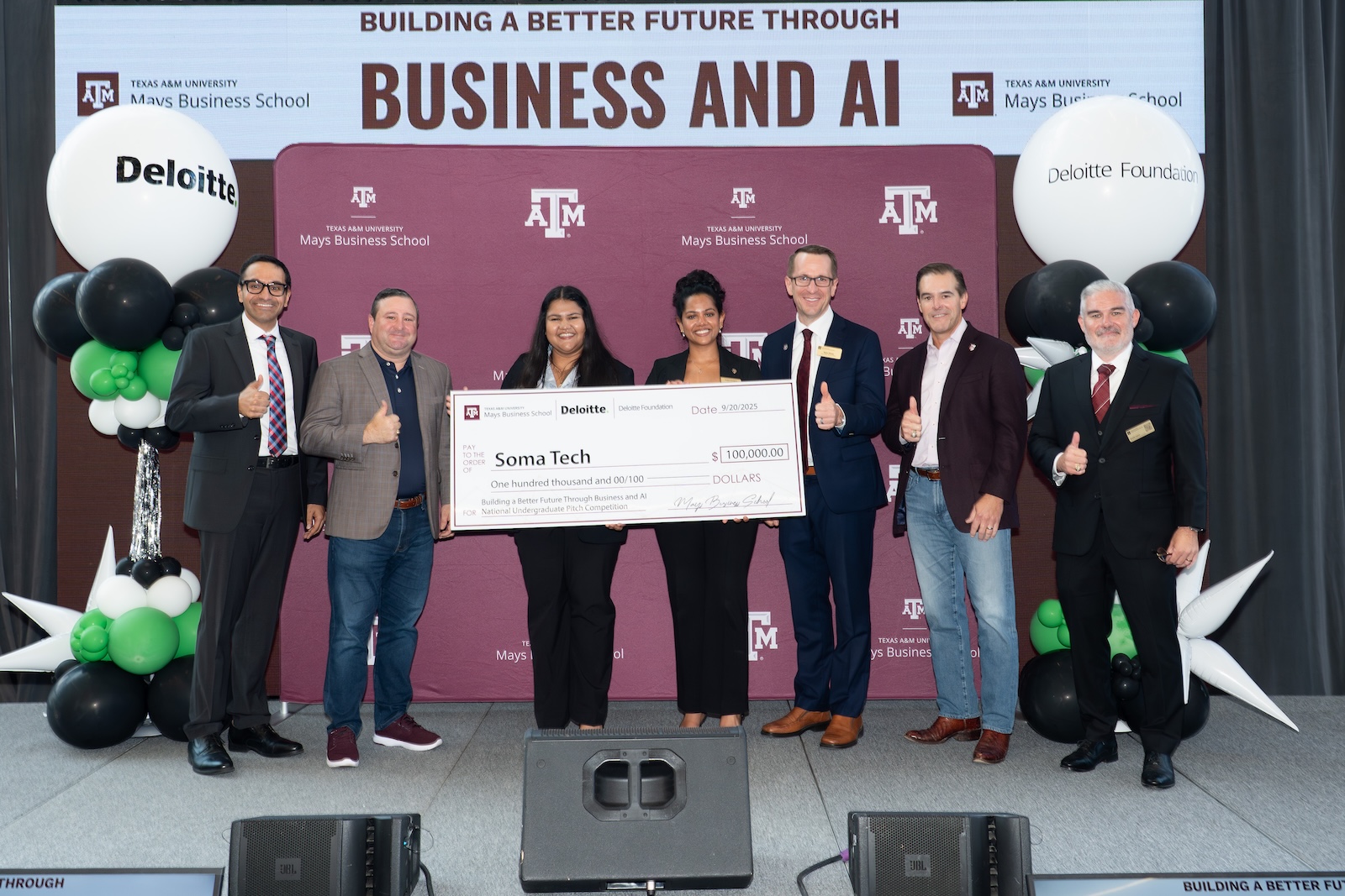 Two women from Soma Tech holding a giant check alongside Mays Business School and Deloitte leaders.
