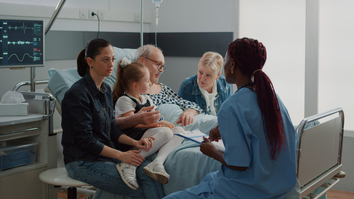 adult-patient-care-1-1408×792 A middle aged patient in a hospital bed surrounded by caring family